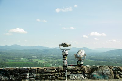 Two coin-operated binoculars covered in stickers on a stone wall overlooking a valley