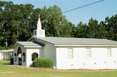 A small white church with a steeple and arched entrance among green trees